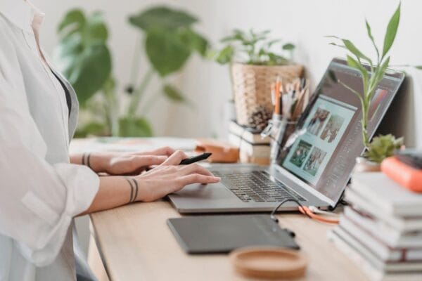 Someone is sitting at a desk, using a laptop with photo editing software open on it. The desk is thoughtfully arranged with plants, a drawing tablet, jars of pens, and a pile of books. Their hands are on the keyboard.