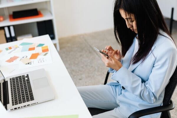 A woman wearing a light blue suit is sitting at a white desk, concentrating on her phone. Beside her are an open laptop and design papers with color samples, showing she is serious about her design work. Behind her, shelves filled with books and folders indicate it's a busy and organized space.