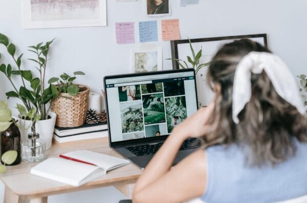 A person wearing a white hair tie is sitting at a wooden desk. There are potted plants, a notebook with a red pen, and photos and notes on the wall. They are looking at pictures on their laptop.