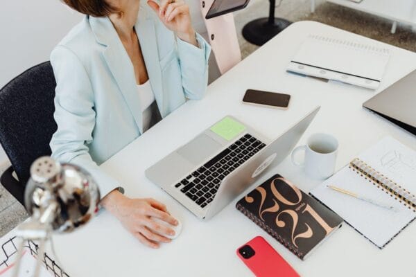 A person is sitting at a white desk with a laptop, coffee cup, smartphone, notepad, and some papers. They are wearing a light-colored blazer and have their hand on their chin as if thinking. Various office supplies are around them. The setup aims to help them work efficiently and stay focused.