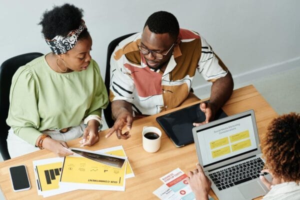 Three people are working together in an office. Two of them are talking about some papers and a tablet on the table, with one of the papers being yellow. The third person is using a laptop and looking at a presentation slide about ways to grow the business. There is also a coffee cup on the table.