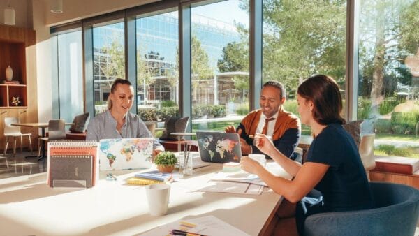 Three coworkers are sitting at a table in a bright, modern office with big windows. Each has a laptop decorated with world maps. They are talking animatedly about growth, with papers and coffee on the table, while sunlight pours in through the windows.