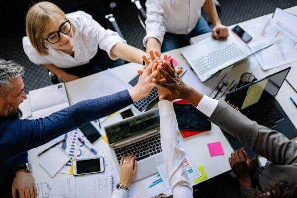 A diverse group of coworkers sit around a table with laptops, papers, and sticky notes. Showing teamwork and working together, they all reach into the center for a group high-five, showing their shared commitment to growing and being involved in the community.