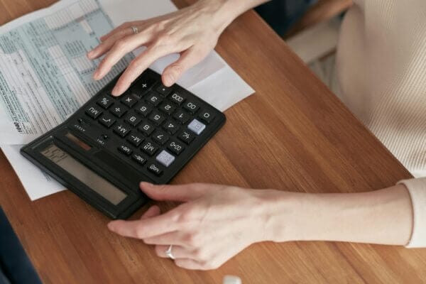 Someone is using a black calculator on a wooden table. There are some papers next to the calculator. The person, wearing a light-colored shirt, is pressing the buttons on the calculator, showing that they are doing some math.