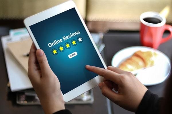 Someone is holding a tablet that shows an "Online Reviews" screen with two stars highlighted, which might be part of an advertisement. There's also a "Submit" button on the screen. In the background on the table, there's a red mug and a plate with a croissant.