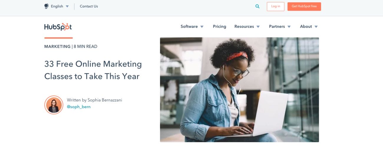 A woman with curly hair and glasses is sitting on some stairs using a laptop. The image has text that says, "33 Free Online Marketing Classes to Take This Year" by Sophia Bernazzani. These classes are part of the digital marketing resources available on the HubSpot website.