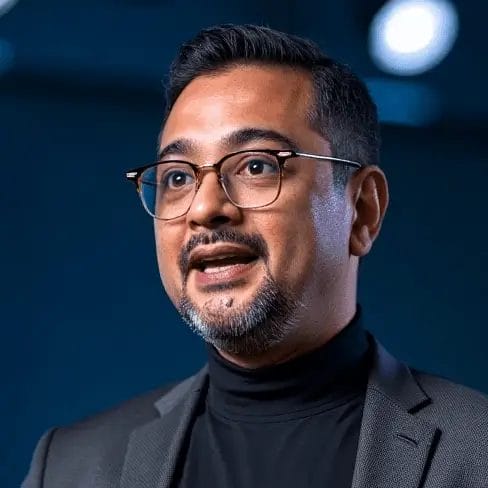 A man who looks like Ruhani Rabin is talking. He has glasses, a neatly trimmed beard, and is wearing a dark suit with a black turtleneck. The background behind him is blurry with round lights.