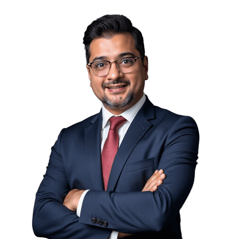 A man wearing a suit, glasses, and a red tie, who looks similar to Ruhani Rabin, is standing in front of a simple blue background. He is smiling and has his arms crossed.