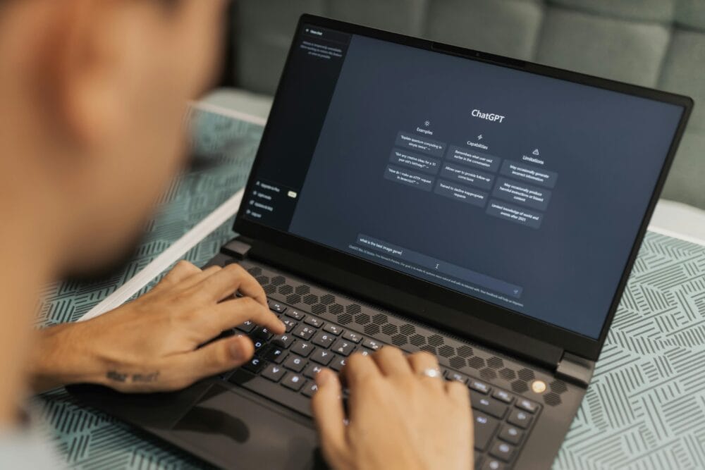 Typing on laptop using ChatGPT, a conversational AI, with options for examples, capabilities, and limitations shown on screen.